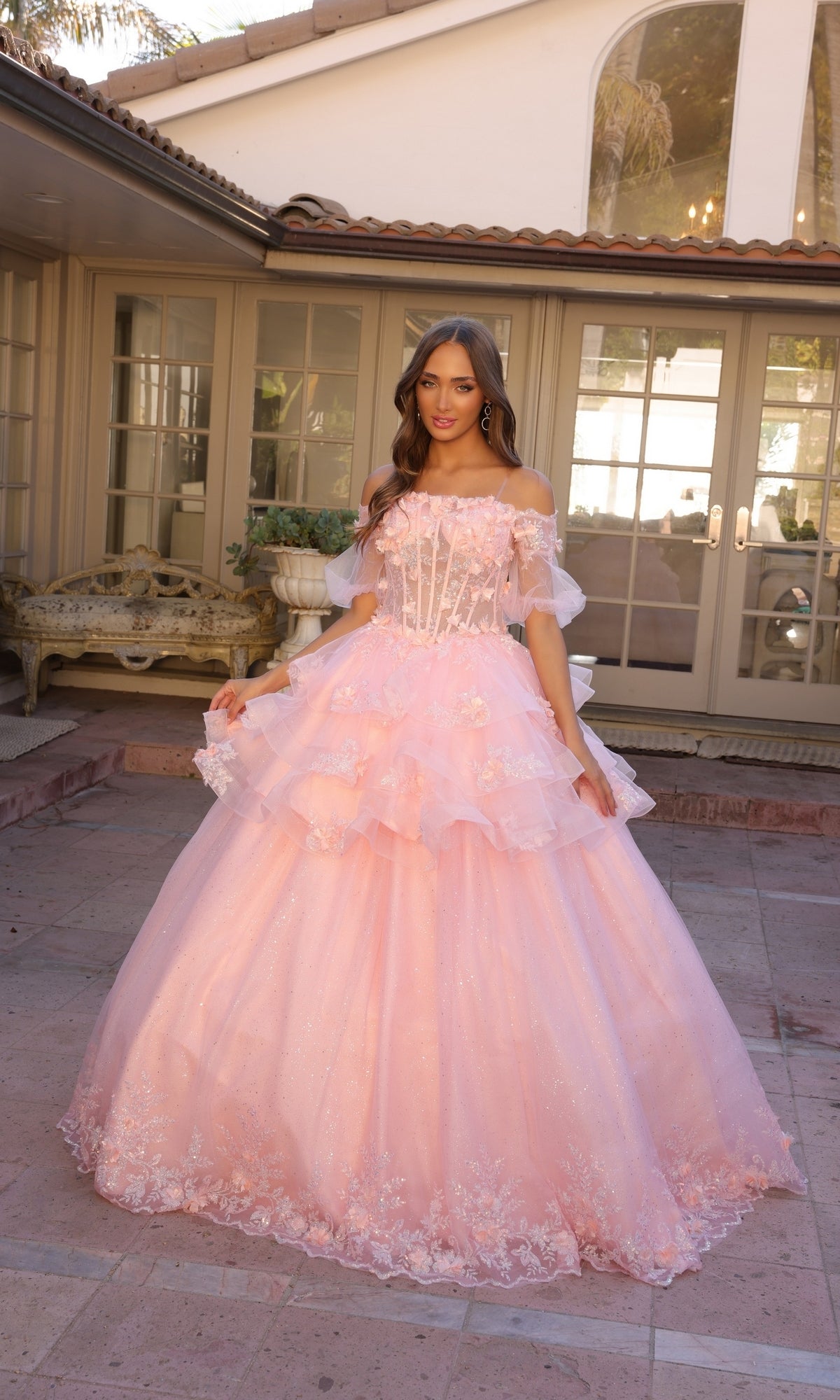 A woman stands outdoors in front of a large windowed building wearing the Nox Anabel Removable-Skirt Formal Ball Gown H1360, a pink off-the-shoulder prom dress with floral details.